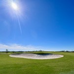 The sun shines brightly above a sand pit surrounded by vibrant greens.